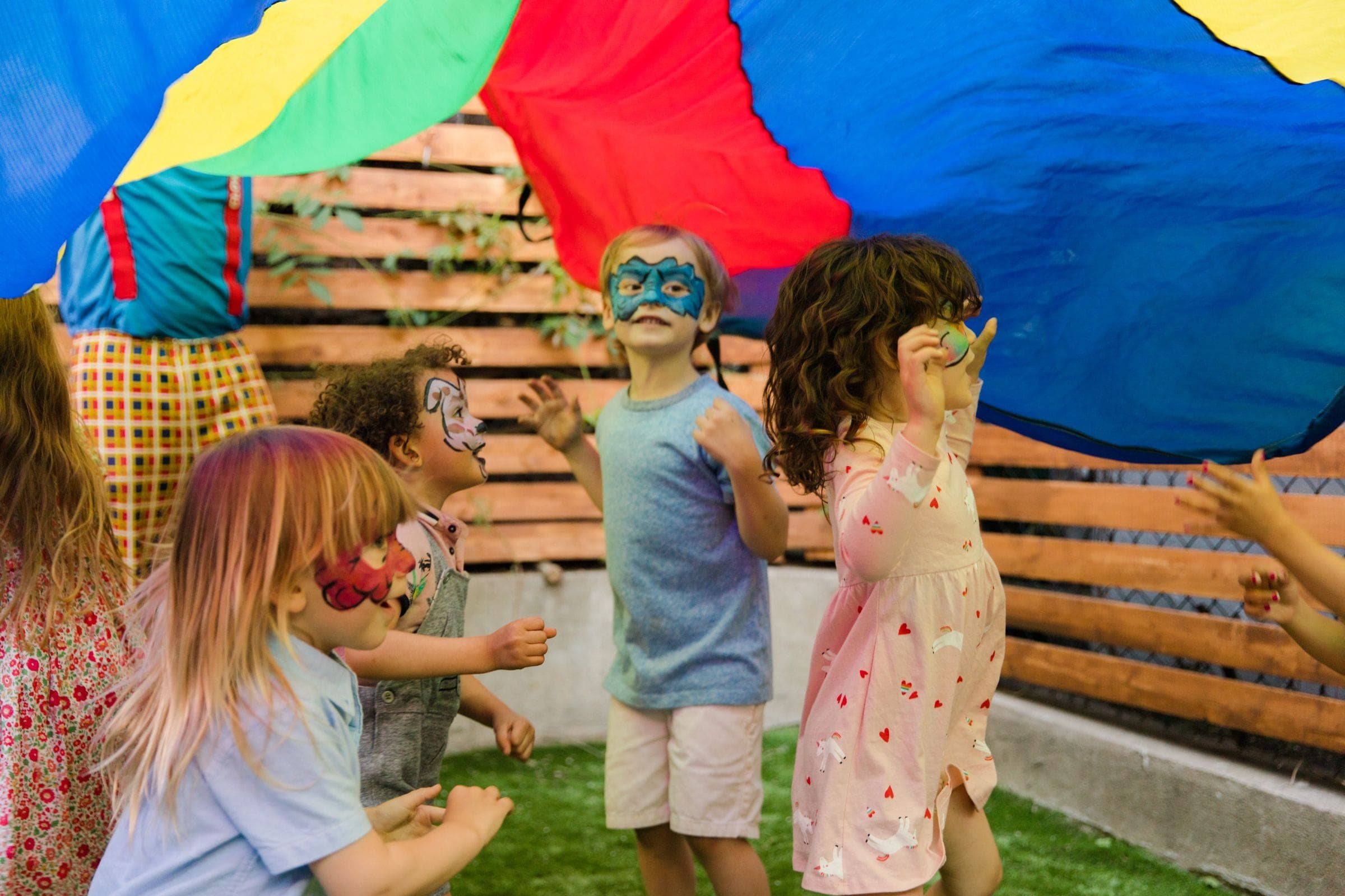 Face-painted children playing together under a rainbow parachute in a garden
