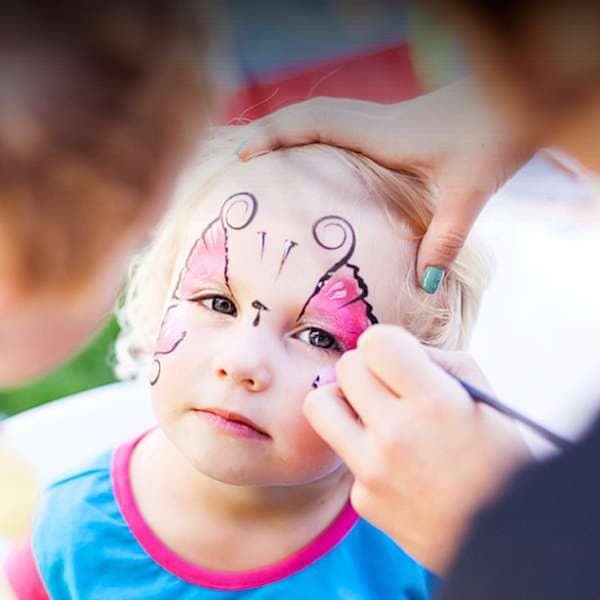 A toddler getting a pink butterfly painted on their cheek outdoors
