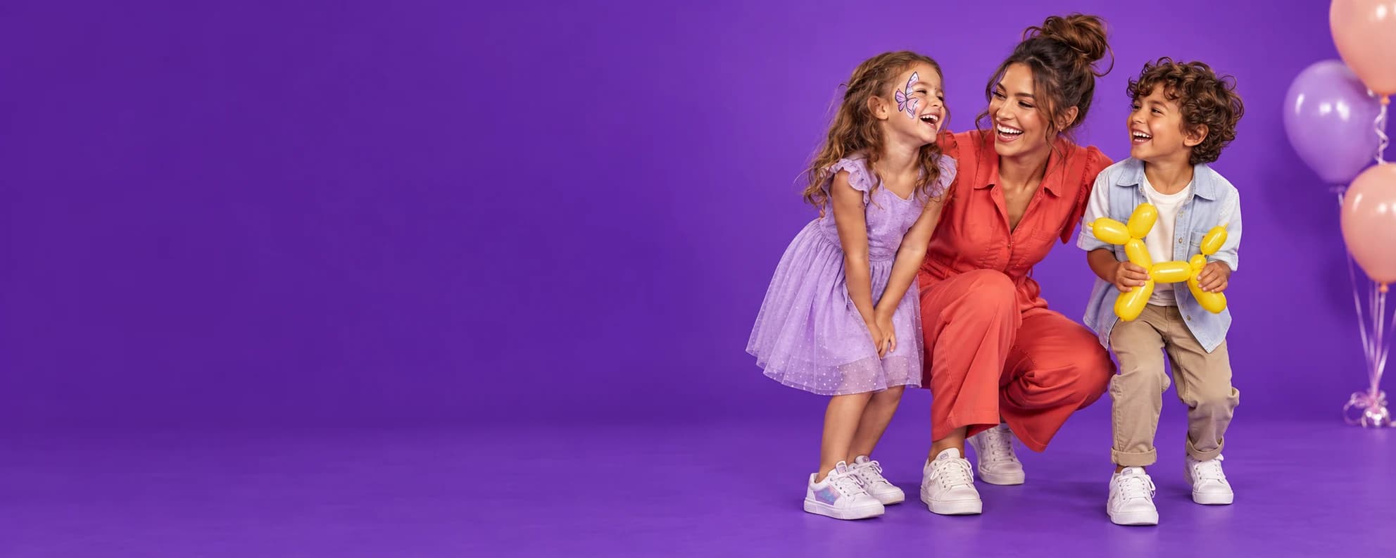 An entertainer laughing with two children, with butterfly face paint and a balloon dog