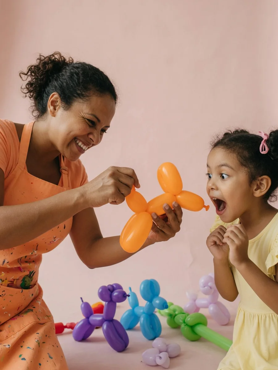 A balloon artist twists an orange balloon dog while a wide-eyed girl watches up close on a blush pink studio backdrop